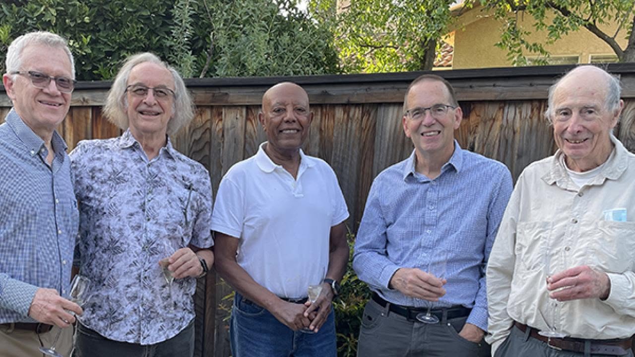 Six NAS members standing side-by-side outdoors by a wooden fence, smiling.
