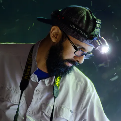 Bearded man in photograph wearing cap-mounted LED headlamp and camera strap, looking down