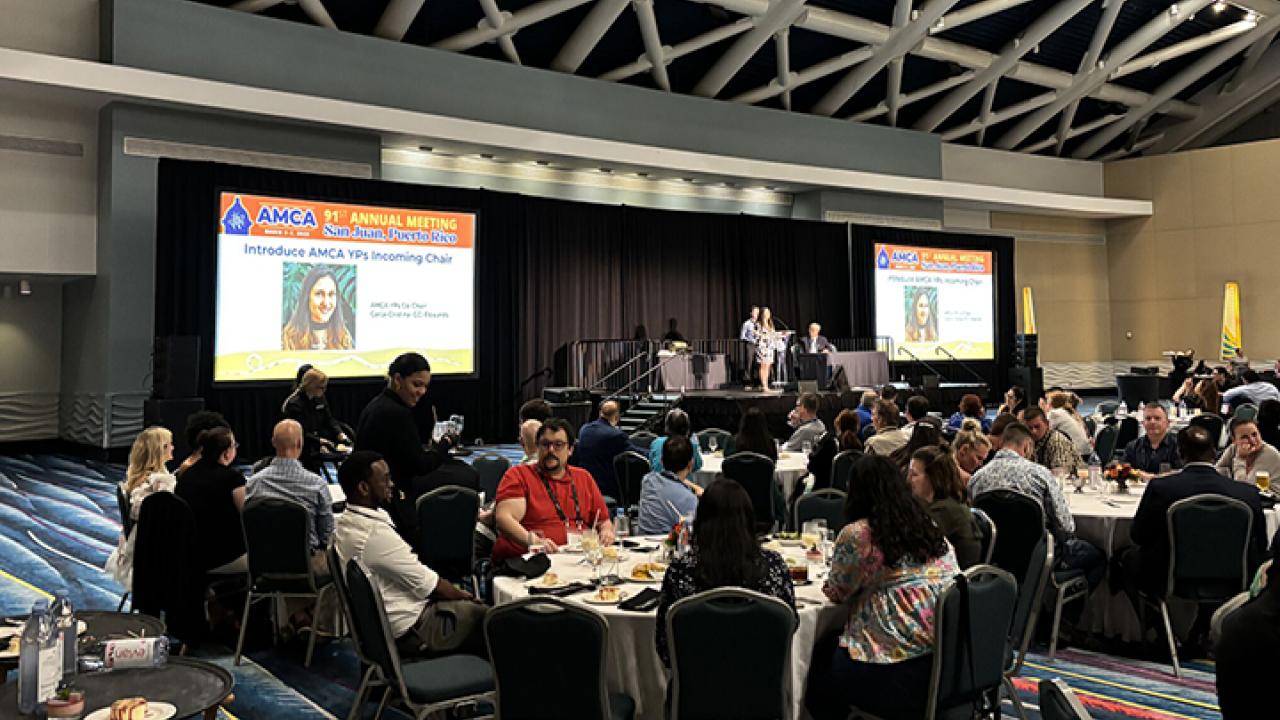 A conference hall with attendees at tables, a stage, and presentation screens.