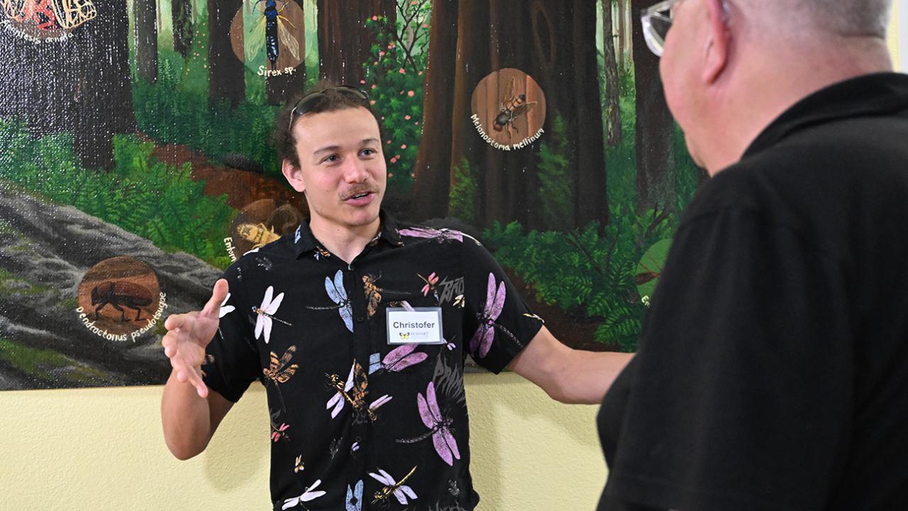 Christofer Brothers in floral shirt gestures while talking to an older bald man in front of forest mural