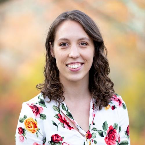 Smiling headshot of postdoctoral fellow with curly brown hair wearing floral blouse, autumn bac