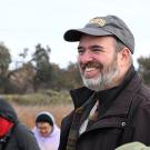 Smiling man with gray beard and cap in dark jacket outdoors with others, cloudy sky