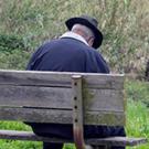 An elderly man, head bowed, sits alone on a bench. 