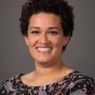 Smiling woman with curly hair wearing a patterned blouse against a gray background.