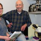Two smiling researchers in a lab with microscopes; woman seated with papers, man standing.Y